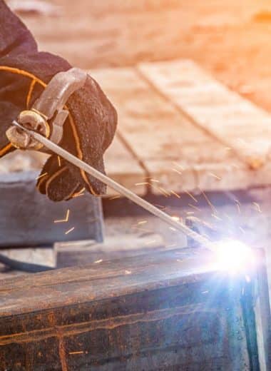 Close up of a young man welder in brown uniform, welding mask and welders leathers, weld metal with a arc welding machine at the construction site, blue sparks fly to the sides